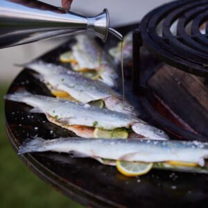 Hand giet olijfolie uit een roestvrijstalen OFYR oliekan over verse visjes met citroenschijfjes op een OFYR plancha-grill.