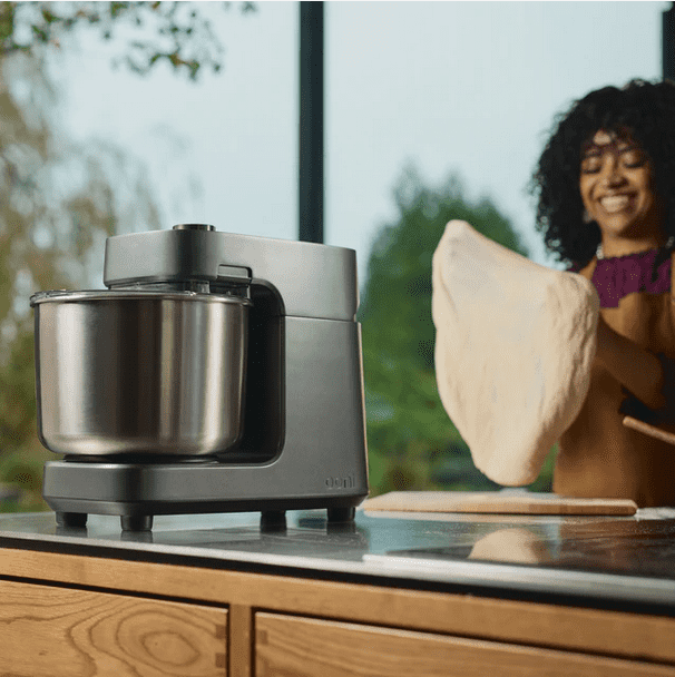 Ooni Halo Pro deegmixer met vers gekneed pizzadeeg Lachende vrouw houdt een grote bal vers gekneed deeg vast naast de Ooni Halo Pro deegmixer met roestvrijstalen kom op het keukenwerkblad.